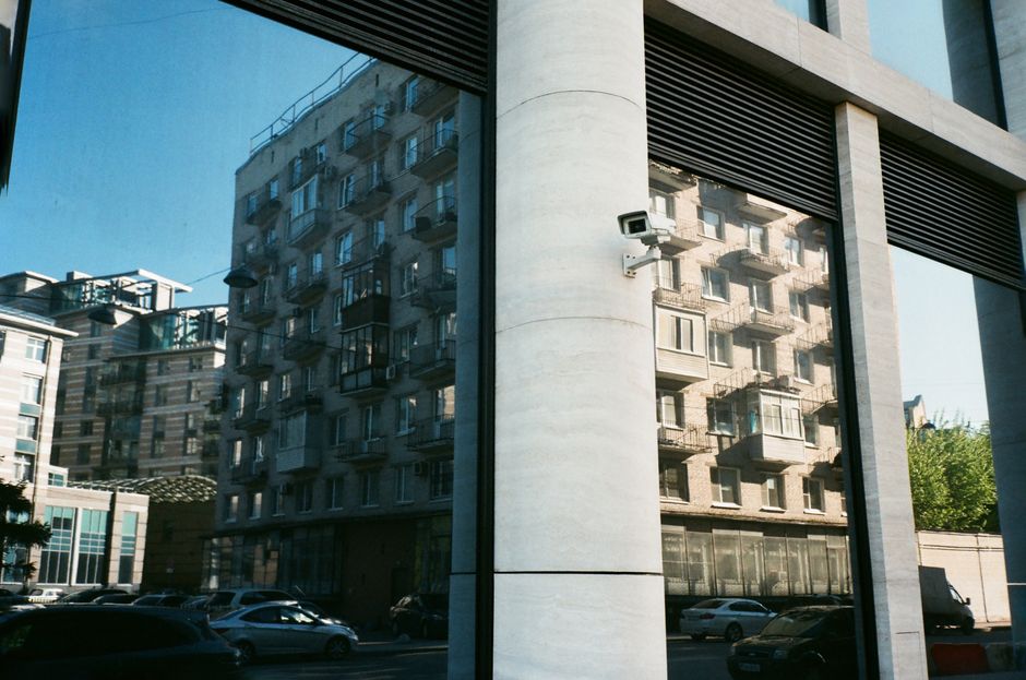 Tall beige multi storey dwelling house reflecting in window of modern construction with small security camera mounted on thick white column