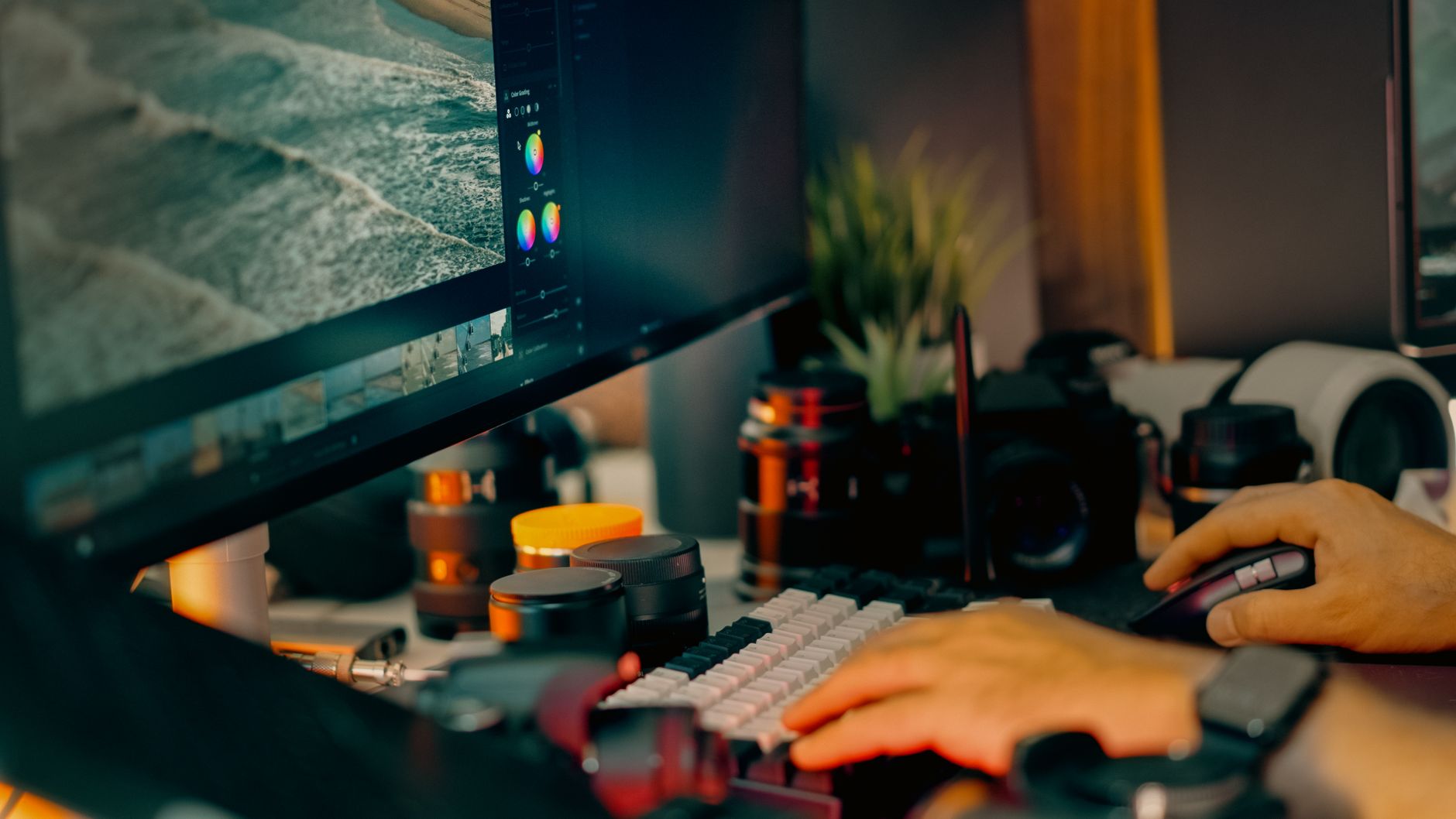 mains sur un clavier et une souris devant un écran affichant un logiciel de retouche photo, objectifs d'appareil photo sur le bureau
